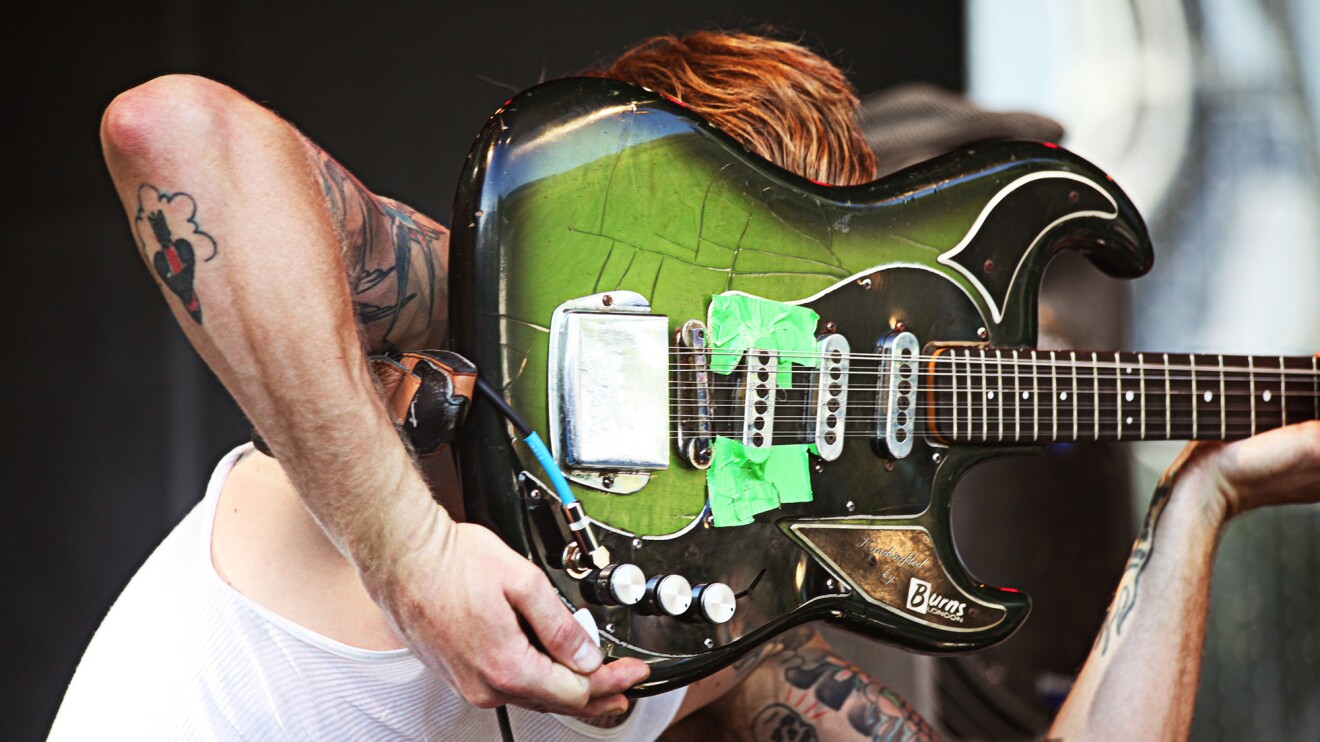 Image of a man hiding his face behind a green and black guitar while performing onstage.