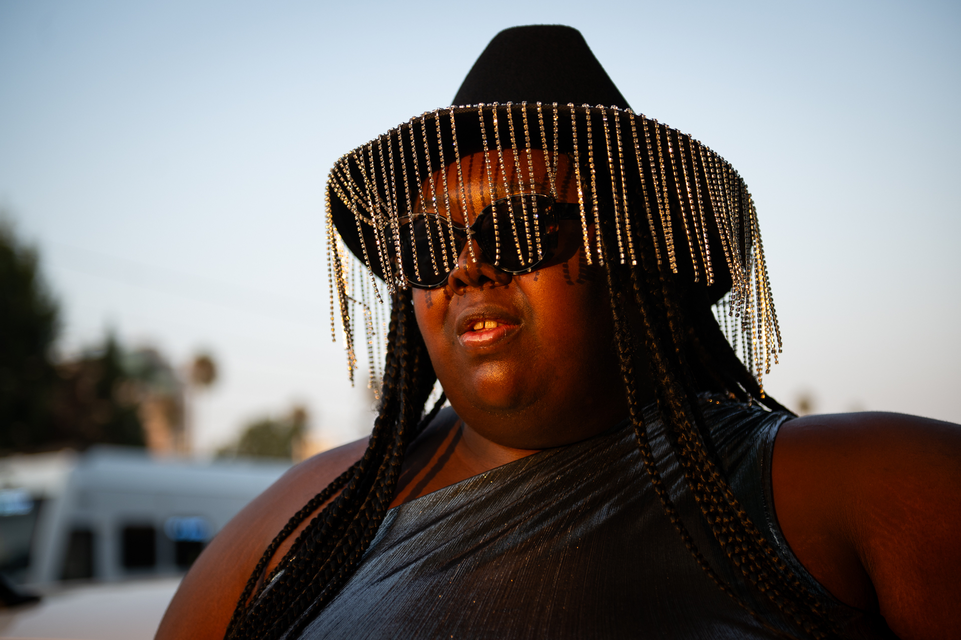 A woman stands in sunset light wearing a black cowboy hat with rhinestone strands hanging around the brim.