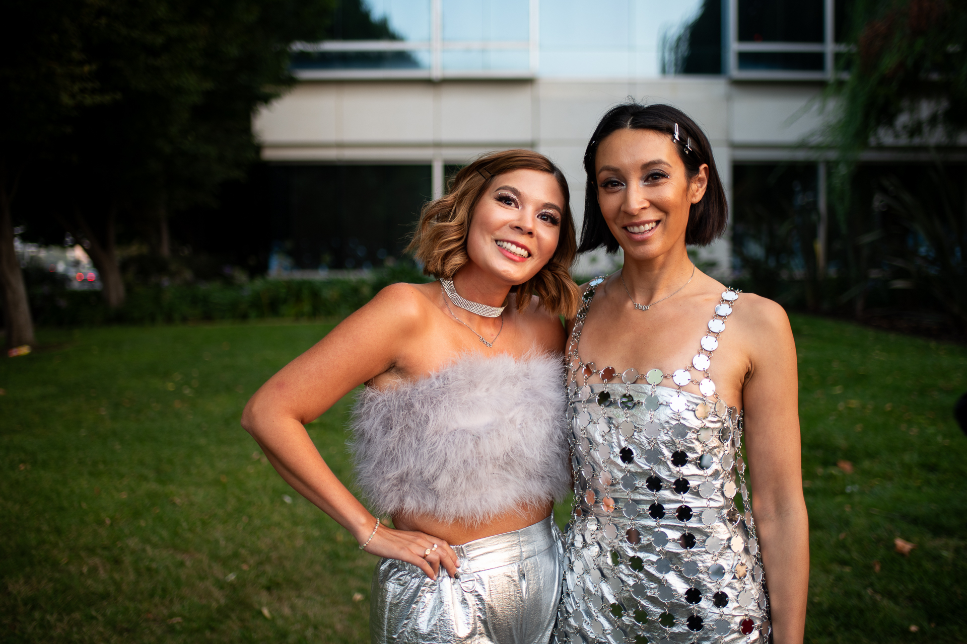 Two women stand with their arms around one another smiling at the camera. The woman on the left wears a feathered top and silver pants, and the woman on the right wears a dress made of small circular mirros.