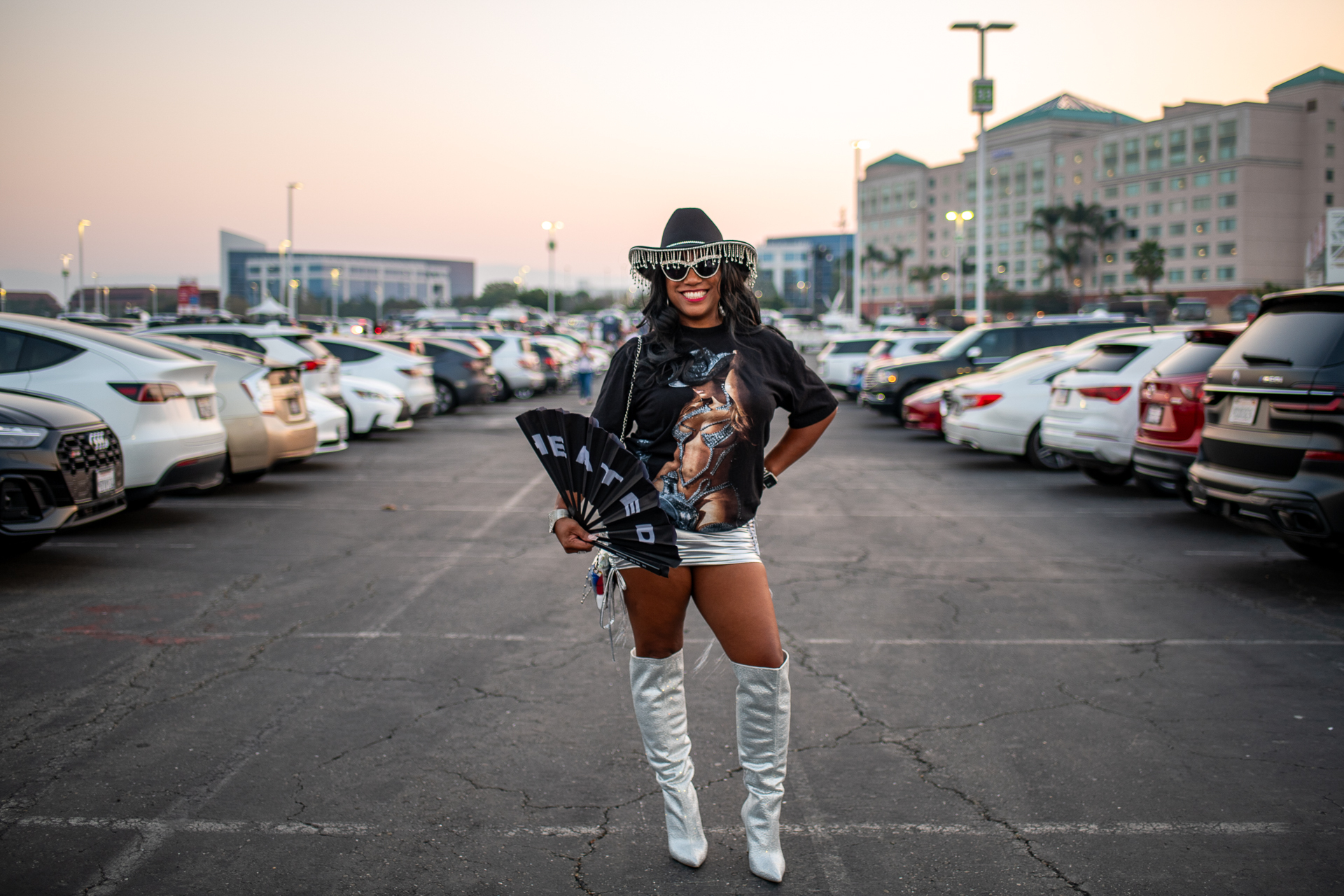 A woman wears a black cowboy hat, Beyonce t-shirt, silver skirt and silver boots with a fan fan while standing in a parking lot.