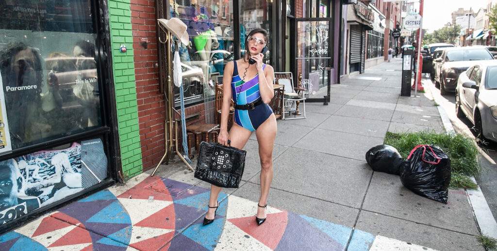 A woman in a belted 80s one-piece bathing suit with a geometric pattern stands on the sidewalk along South Street in Philadelphia. He holds an old-fashioned telephone to her ear, and is wearing large sunglasses, black heels, and is carrying a large black handbag.