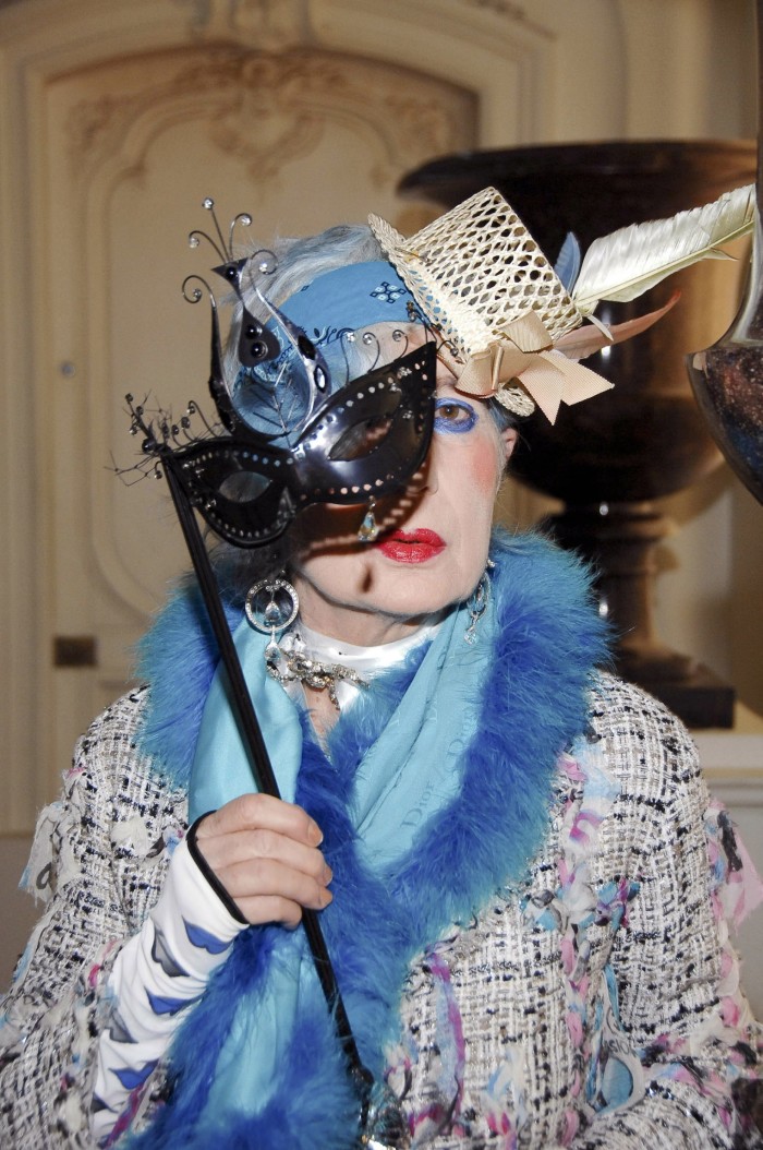 A woman in a feathered hat and blue fringed scarf holds a mask up to her face