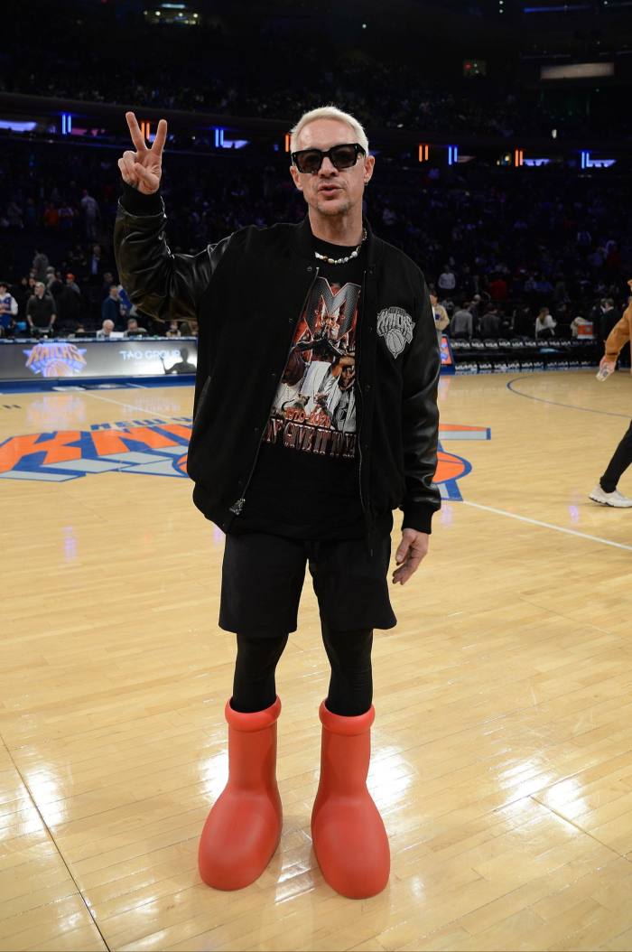 A man in black shorts, black jacket, sunglasses and huge boots stands on a basketball pitch and gives the peace sign 