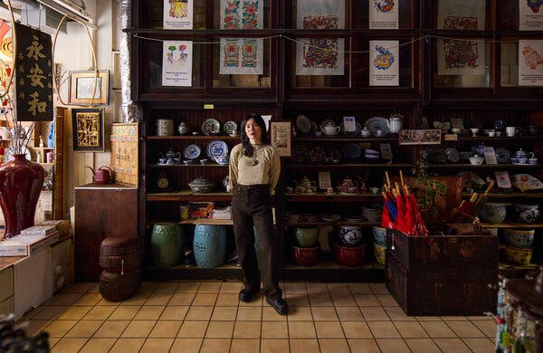 Mei Lum, wearing a long beige T-shirt and dark brown pants, stands in a shop with wooden shelving stacked with ceramics, including stools, plates and platters, bowls and teapots. Prints and calligraphy are hung about.