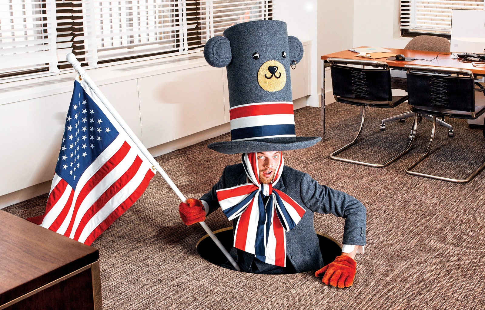 A model dressed in red white and blue and holding an American flag emerges from a hole in the floor of an office.