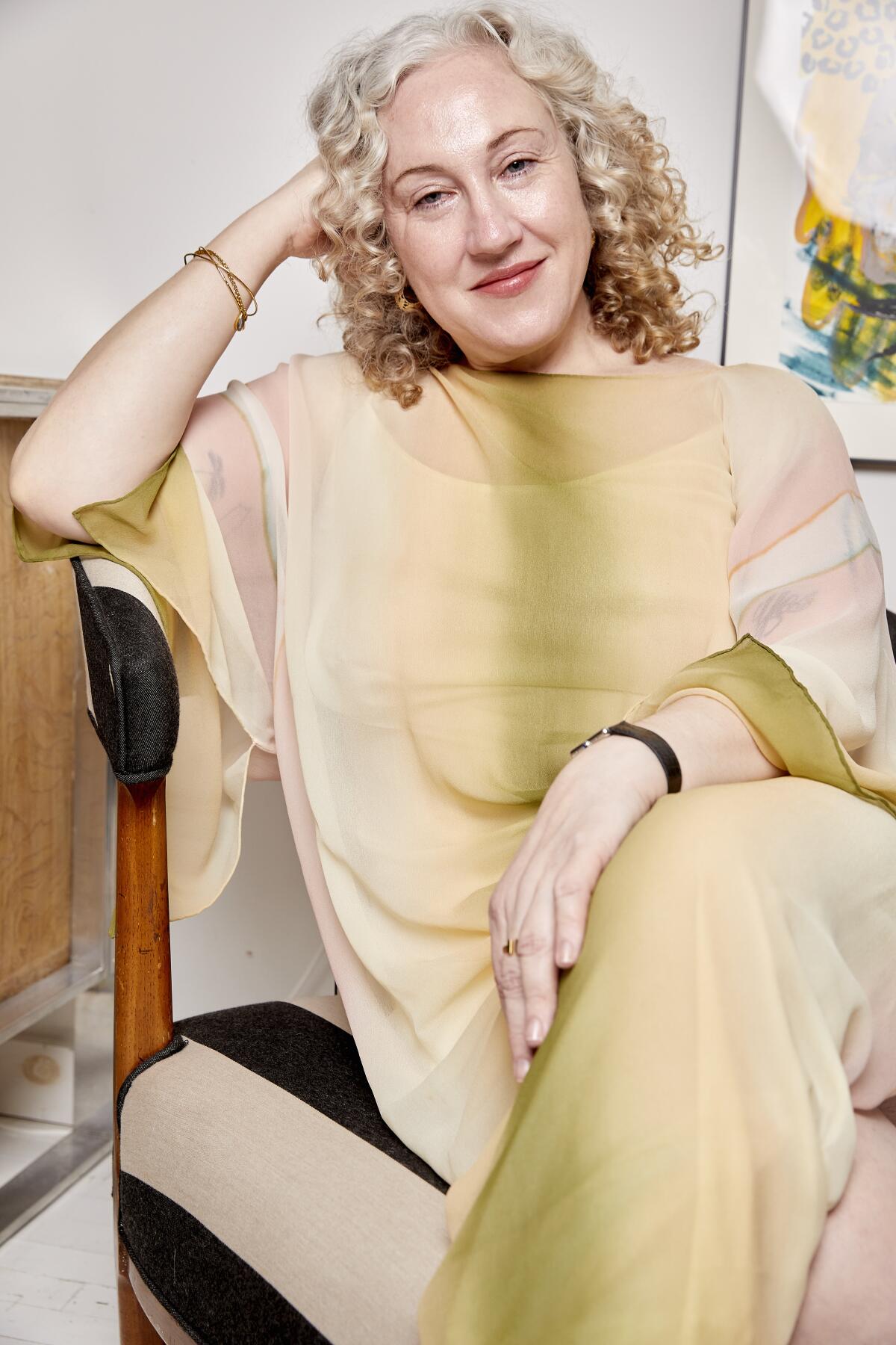 A woman with wavy blond hair seated comfortably in a loose dress of light earth tones.