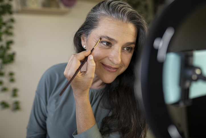 Mature woman filling in eyebrows with eyeshadow and brush.