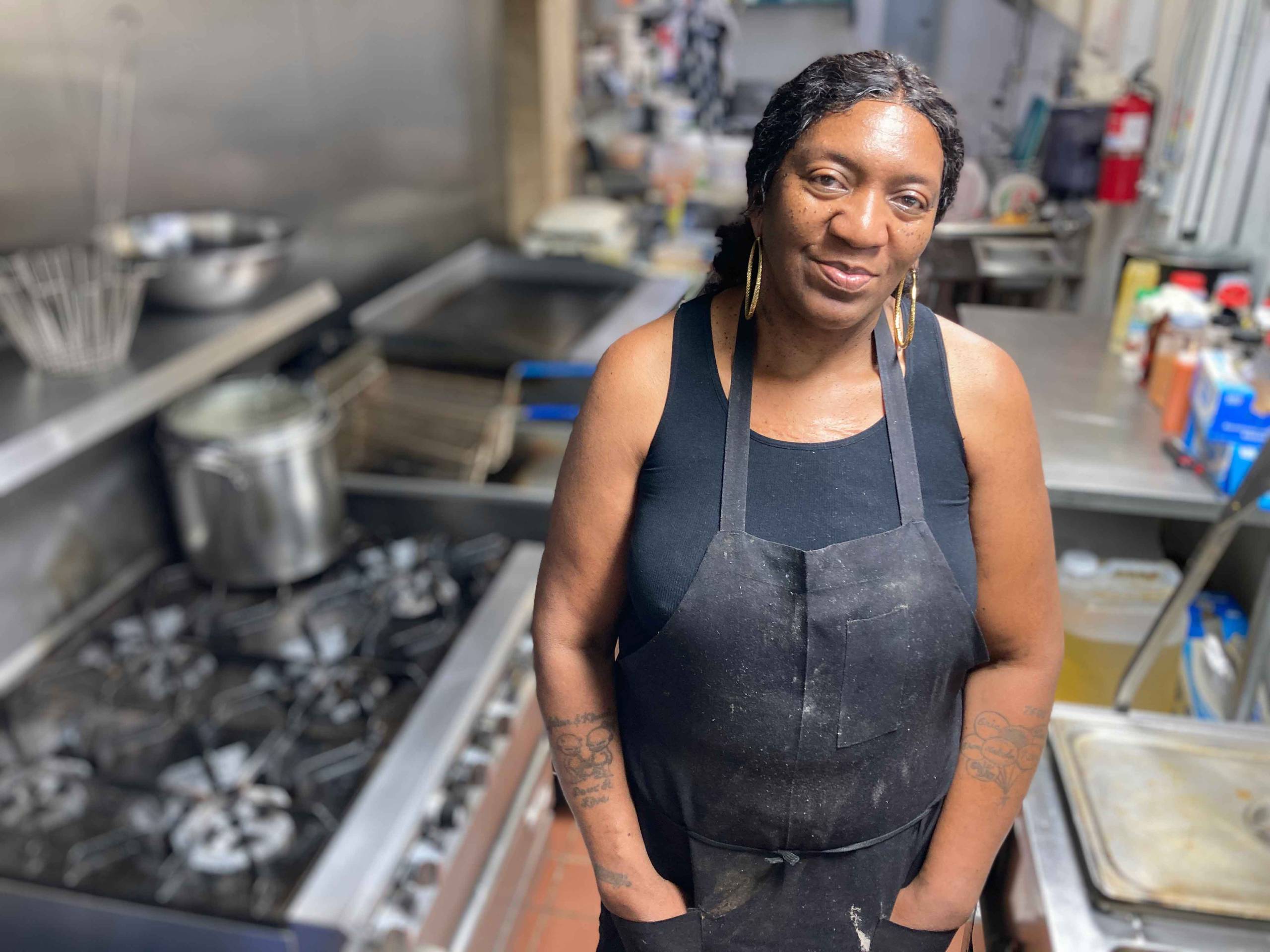 A Black woman in tank top and black apron and gold hoop earrings poses in an industrial kitchen, next to a range.