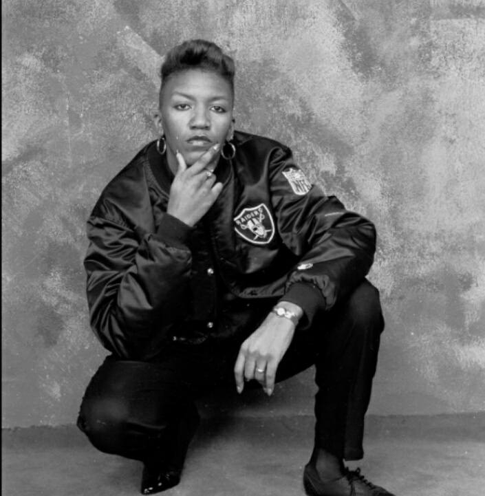 A Black woman in short hair and a Raiders starter jacket and watch crouches in a photo studio.