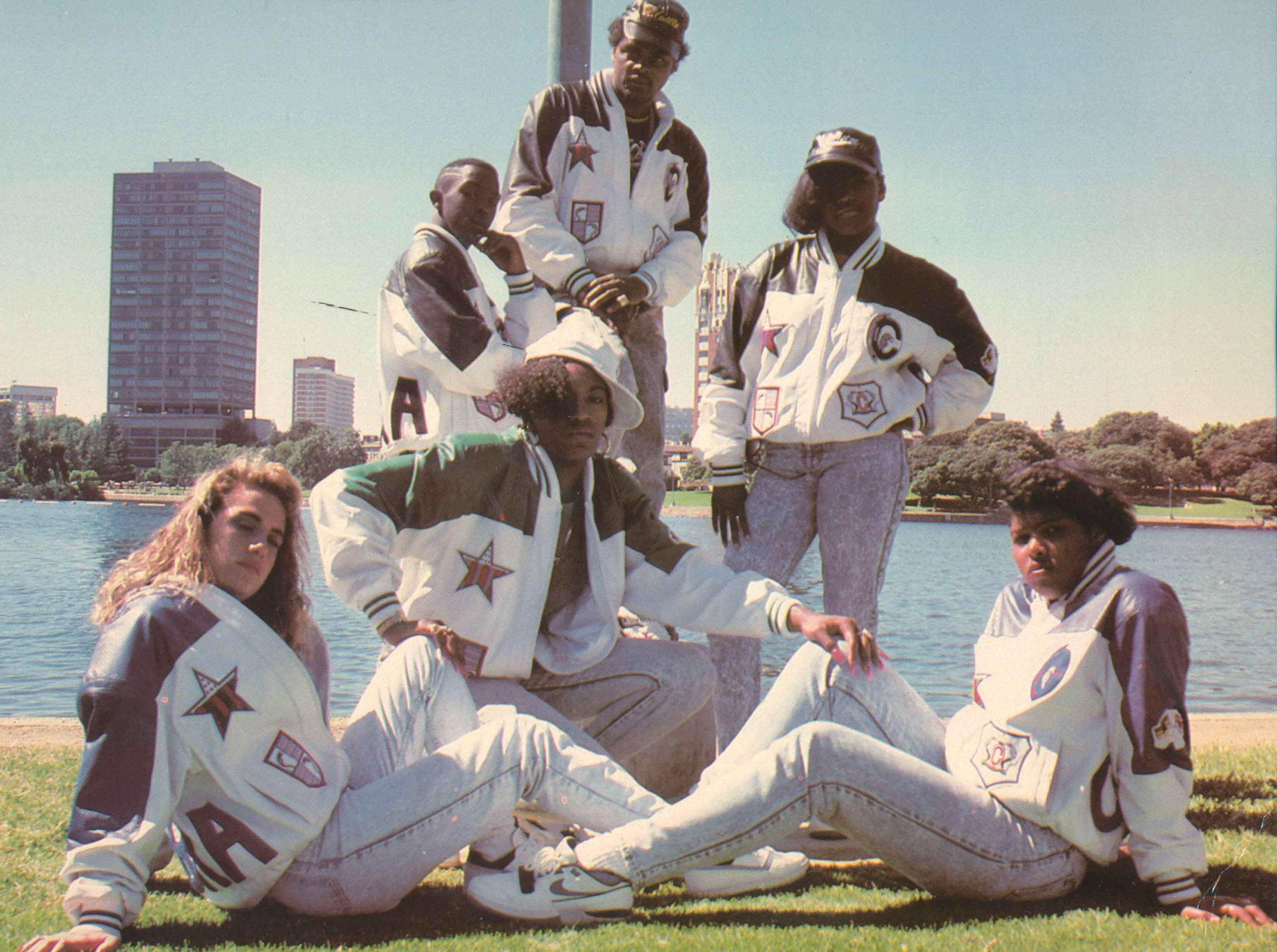 A group of Black teenagers in large jackets and faded jeans pose in front of a lake.