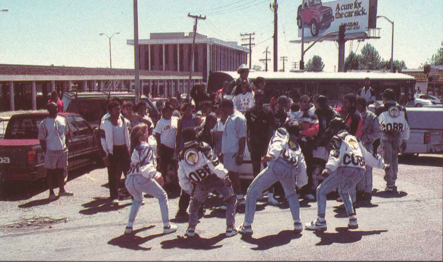A group of Black teenagers in large jackets and faded jeans dance in a parking lot.
