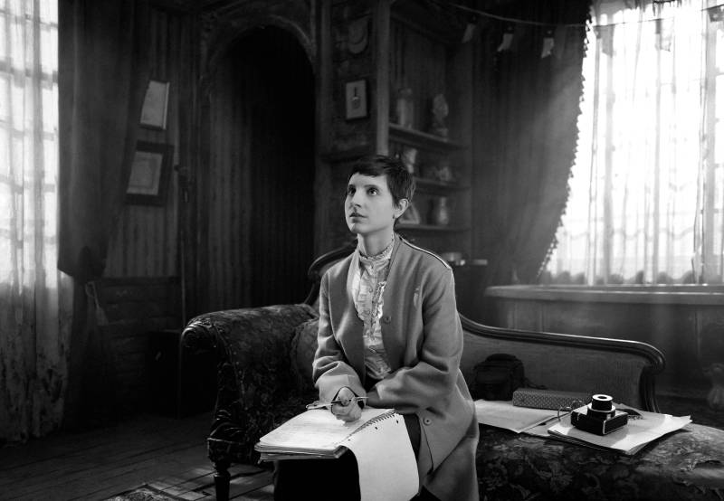 a black and white still image from a film of a young woman in a long cardigan holding a notebook as she sits on a bench, looking up