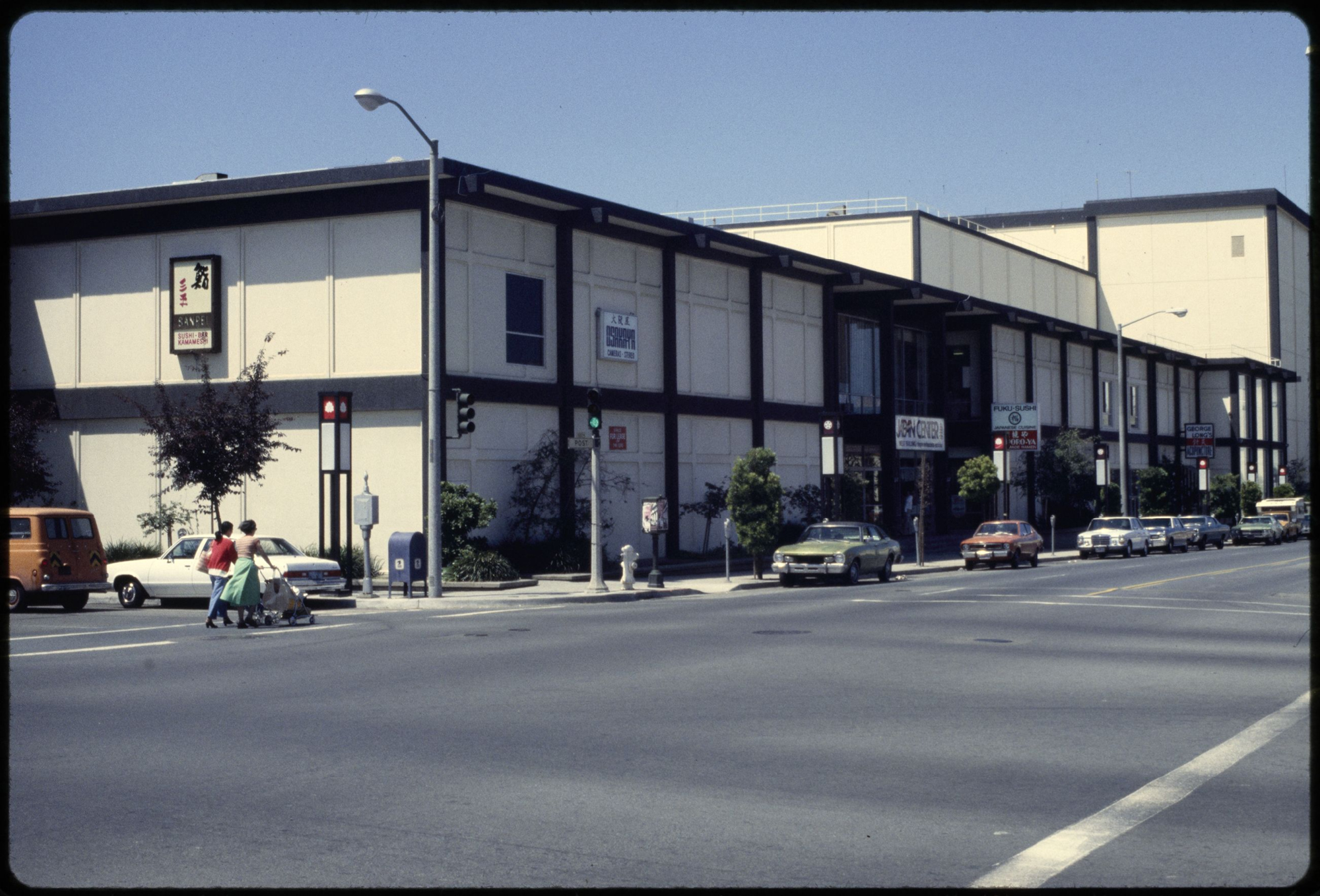 Color photograph of long two-story white building with dark trim and cars parked along curb