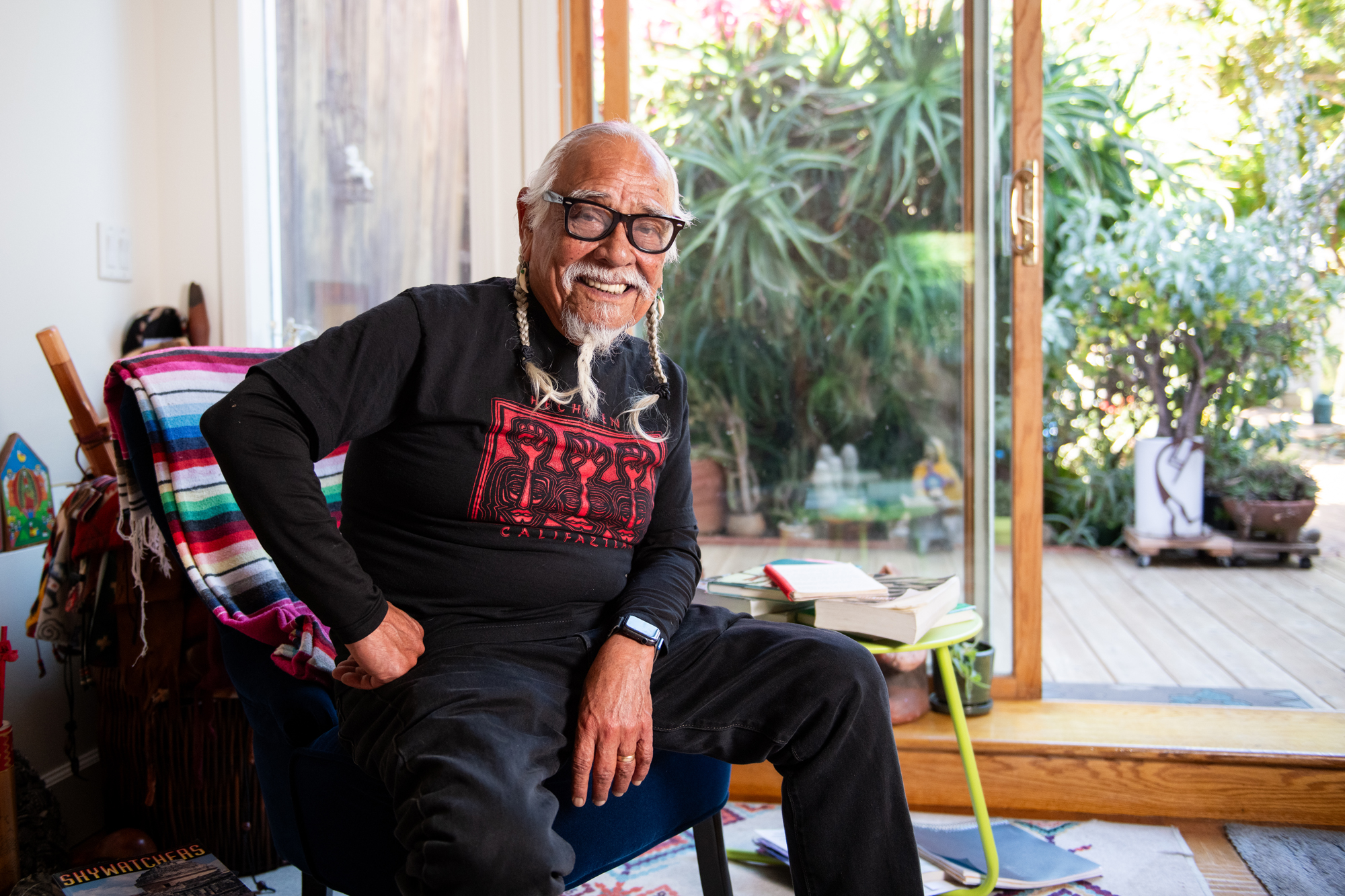 An older man with white braids and glasses sits in a chair smiling, with one arm propped on his hip