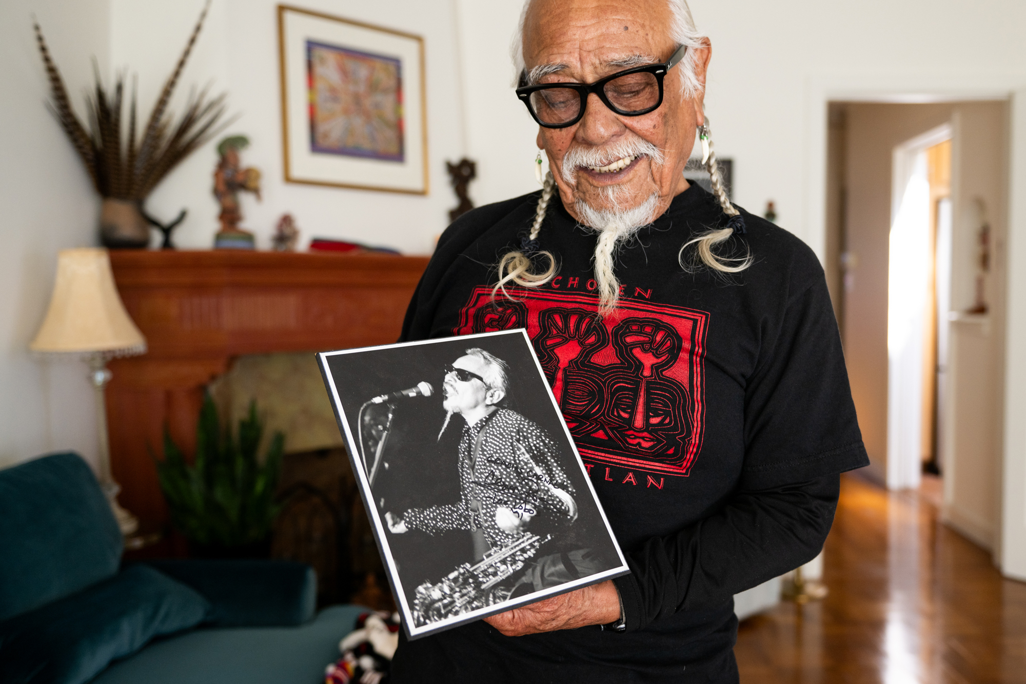 An older man with white braids and glasses holds a black-and-white picture of himself in sunglasses singing into a mic with a guitar hanging in front of him