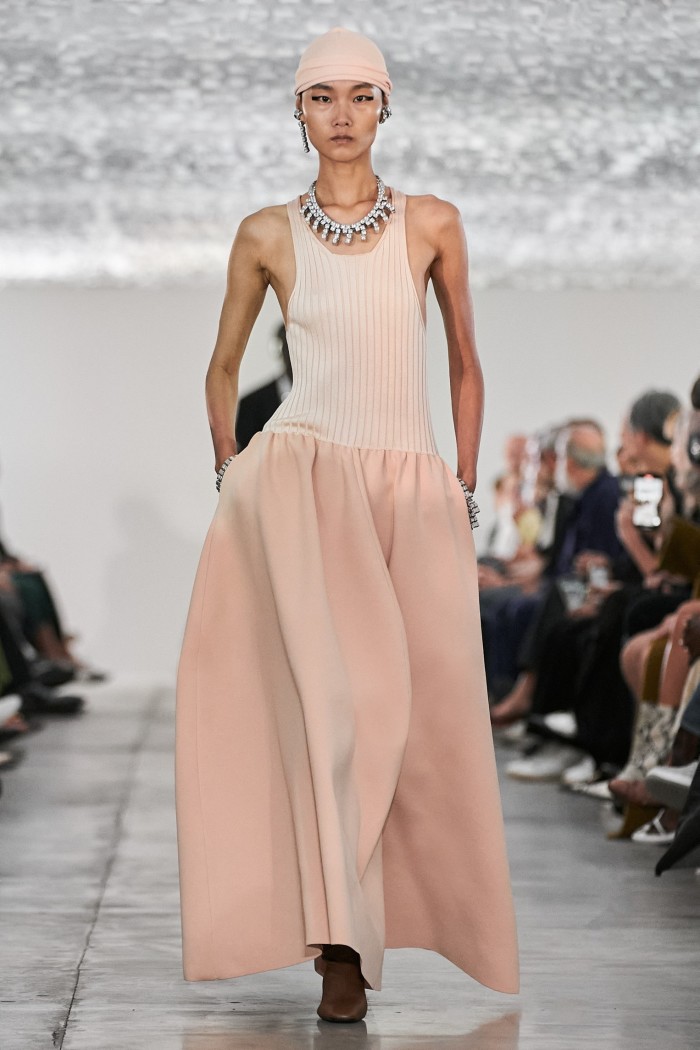 A female catwalk model in a sleeveless dress of peachy pink, with full-length sweeping skirts — plus a tight-fitting cap of the same colour