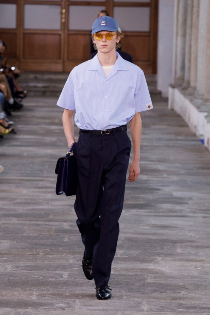 A male model on a paved catwalk wearing a royal blue baseball cap, pale blue short-sleeve shirt and plain black trousers and shoes