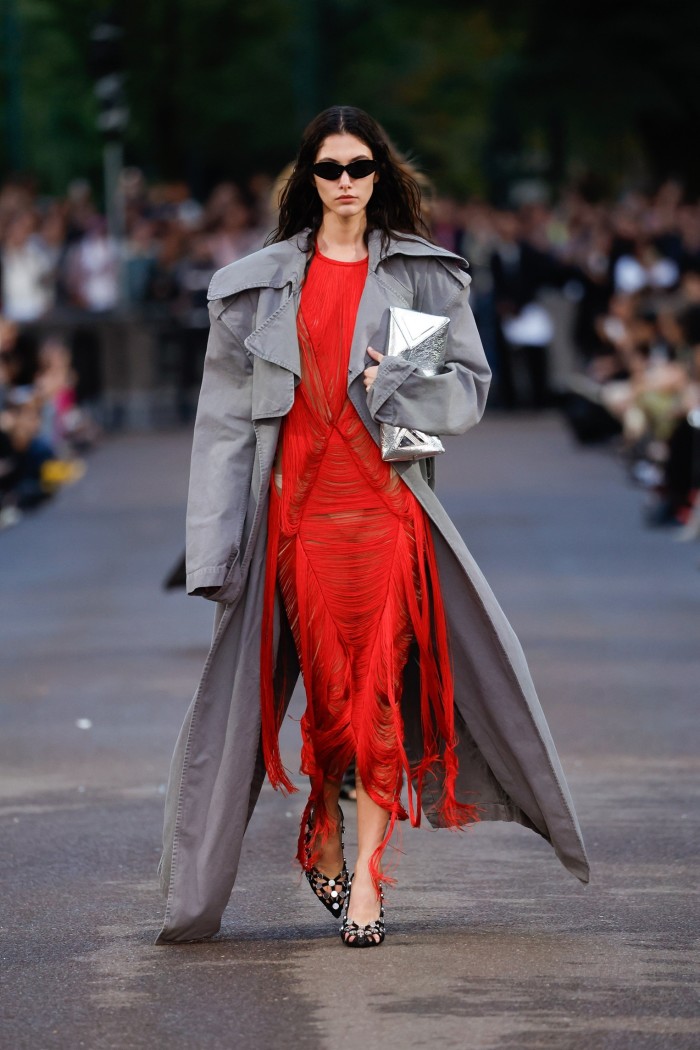 A catwalk model at an outdoor fashion show wearing a wide, loose grey trenchcoat over a red dress