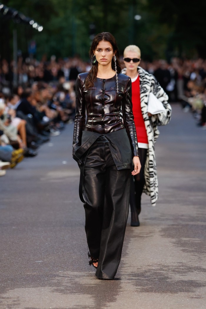 A catwalk model at an outdoor fashion show wearing a tight glossy black top and black leather-look flared trousers