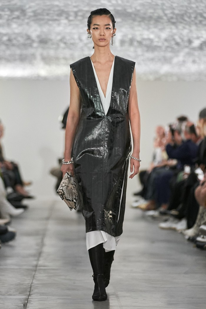 A female catwalk model in a glossy, sleeveless black dress that falls below the knee and overlaid on white cotton