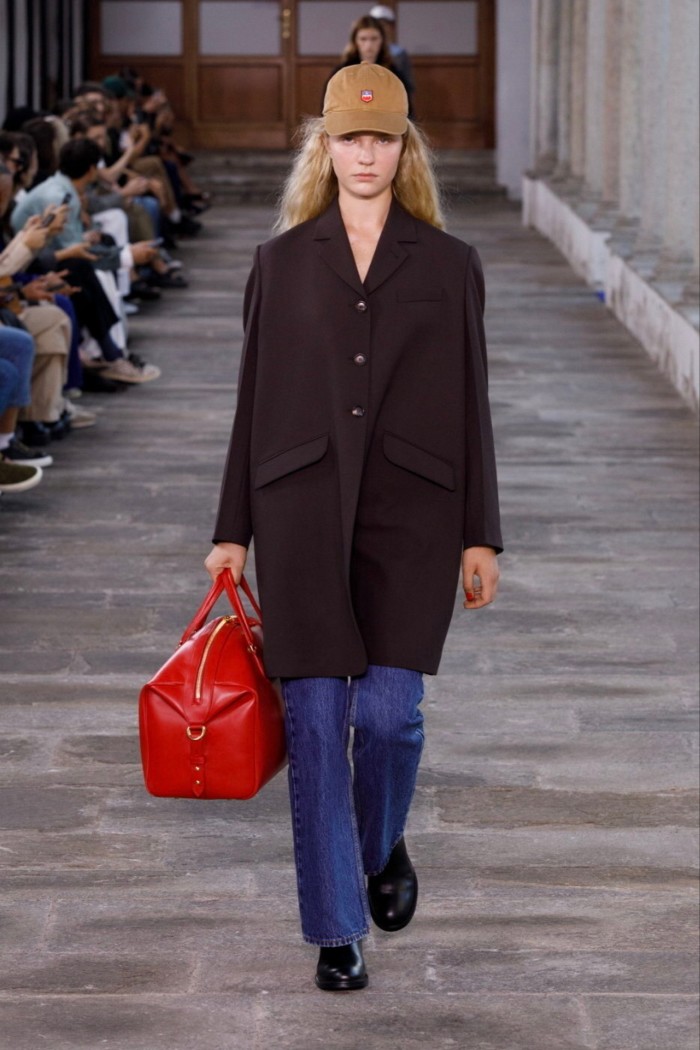 A female fashion model on a paved catwalk, wearing jeans, a caramel-coloured baseball cap, wide chocolate-brown coat and carrying a red bag 