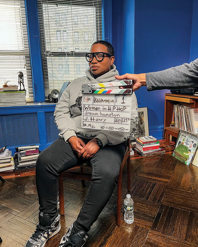 photo of seated short-haired woman with large glasses by bookcases in room painted electric blue with arm reaching in front holding film clapboard for Netflix documentary