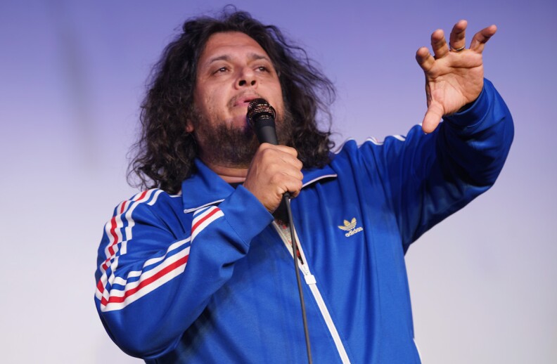 Comic Felipe Esparza holds a mic while outstretching one hand while wearing an Adidas track suit.