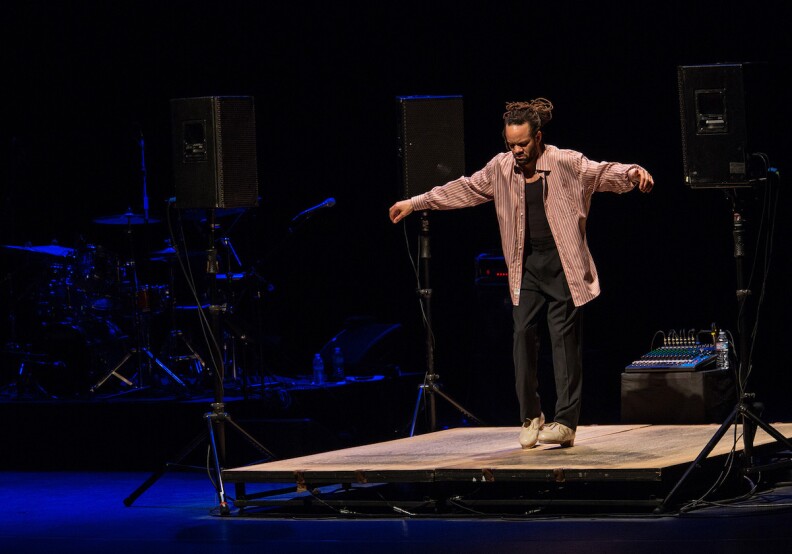 Tap dancer Savion Glover performs on a small miked stage along with a drum kit seen in the dark to our left. 