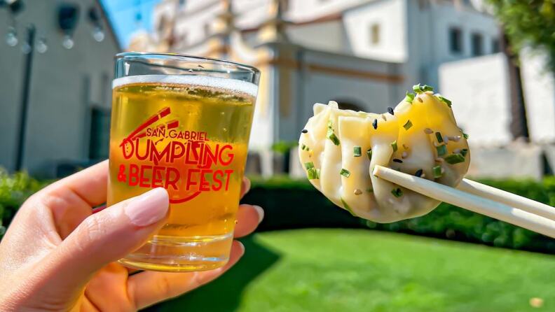 A manicured hand holds a small beer at left and chopsticks hold a single dumpling against a backdrop of a mission church in the daylight for the San Gabriel Dumpling and Beer Fest.