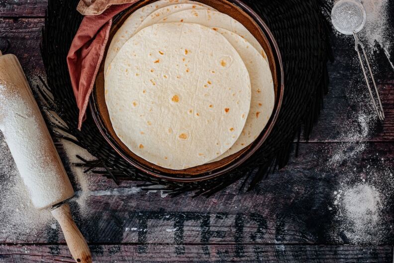 A stack of tortillas sit in a pot with a rolling pin at left and flour on the wooden surface. The photo is taken from above. 