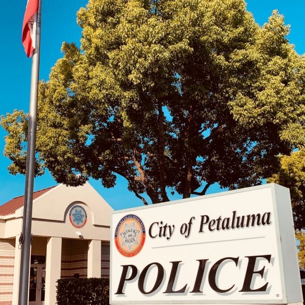 Close up of a sigh that reads City of Petaluma Police. A building with a badge included above its entrance is in the background