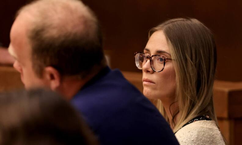 A white woman in tortoiseshell glasses looks across a room. A white man next to her is out of focus.