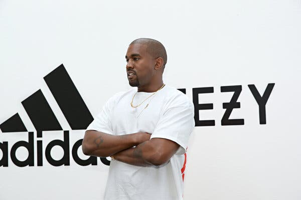 Kanye West, wearing a white T-shirt and crossing his arms, stands in front of a backdrop that says “Adidas” and “Yeezy.”