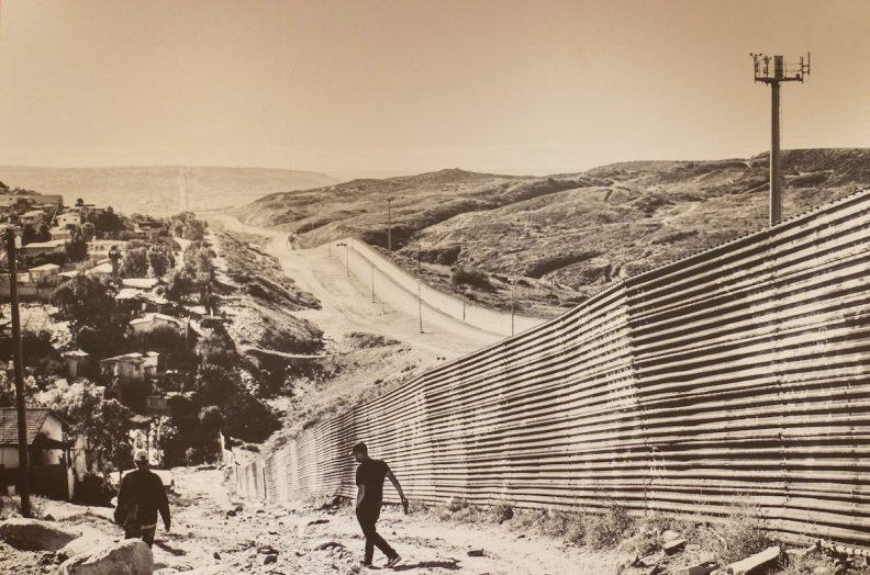 Photograph taken at the US/Mexico border by AMBOS project photographer Gina Clyne. The image is in sepia tone with two figures in the foreground walking along a long wall. 