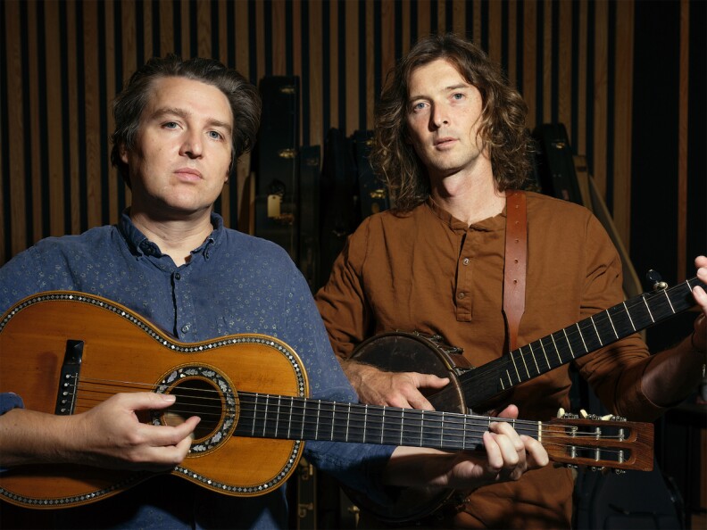 One man holds and plays a guitar at left and the other man at right holds an old banjo. Both face the camera in this promo shot for The Milk Carton Kids.