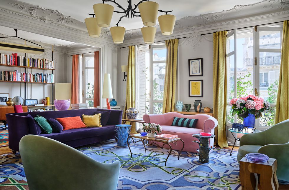 living room with three floor to ceiling windows with yellow and orange drapes, two sofas, two armchairs, a curved leg cocktail table, framed artworks, three artful side tables, vases with flowers, blue patterned rug