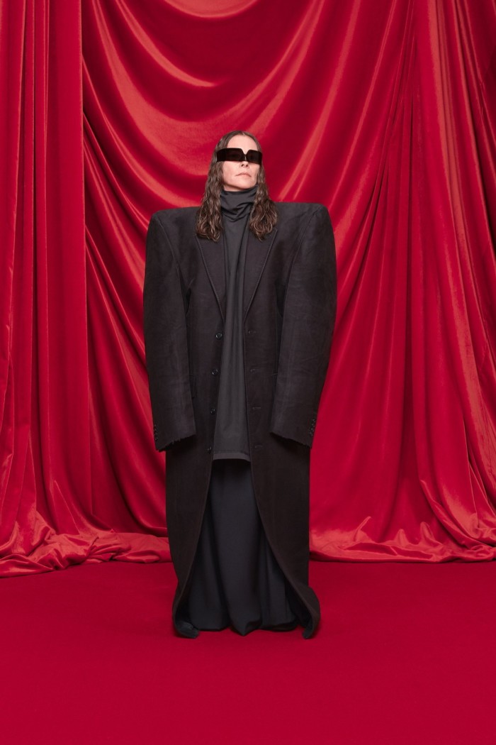 A woman stands in front of red curtains wearing an extremely oversized black coat and some sunglasses