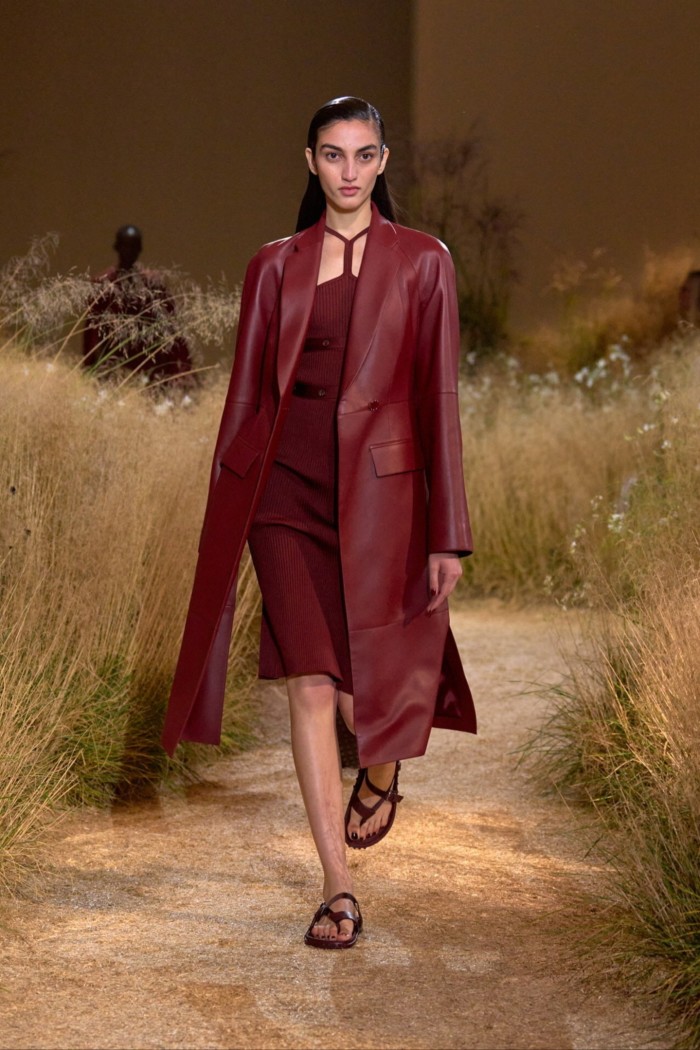 A model walks between grass wearing a burgundy red leather coat over a red dress