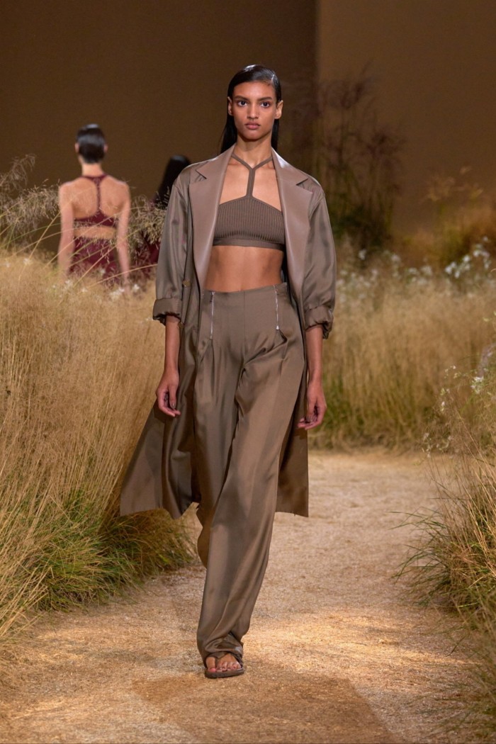 A model walks through grass wearing a lightweight taupe coat over gauzy trousers and bra top