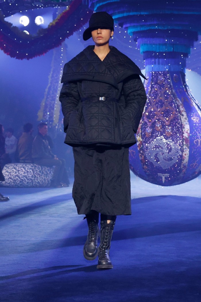 A model walks on a futuristic looking catwalk, wearing black boots, black puffer jacket and baseball cap