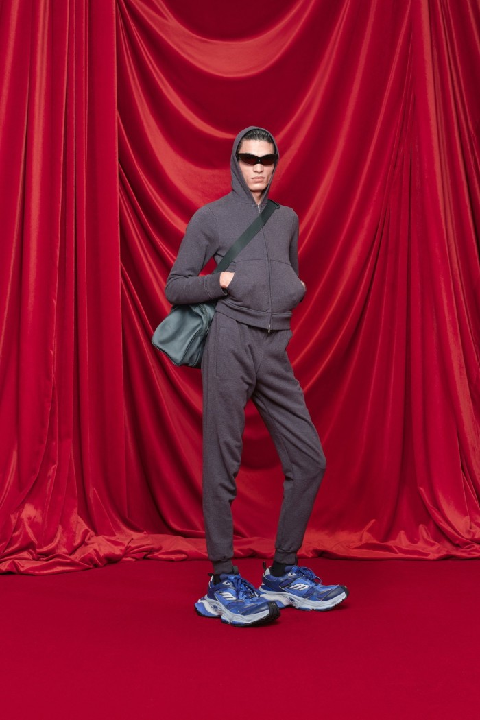 A model stands in front of red drapes wearing tracksuit trousers, hoodie and oversized sneakers