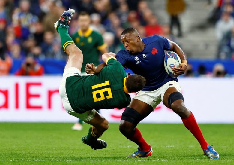 A rugby player in a blue jersey and carrying the ball upends a player in a green jersey.