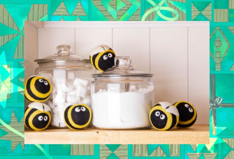 The scene is a laundry room. Large glass jars contain laundry cleaning powders. And surrounding the jars are round woolen balls made to look like bumble bees with yellow and black stripes, while wings, and kooky eyes that look a bit dizzy after a few loads of dryer time. The image is set against a decorative green background accented with stars, gift boxes and strands of ribbon.