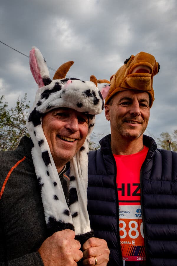 A man wearing a furry cow hat smiles while standing with a man wearing a furry horse hat.