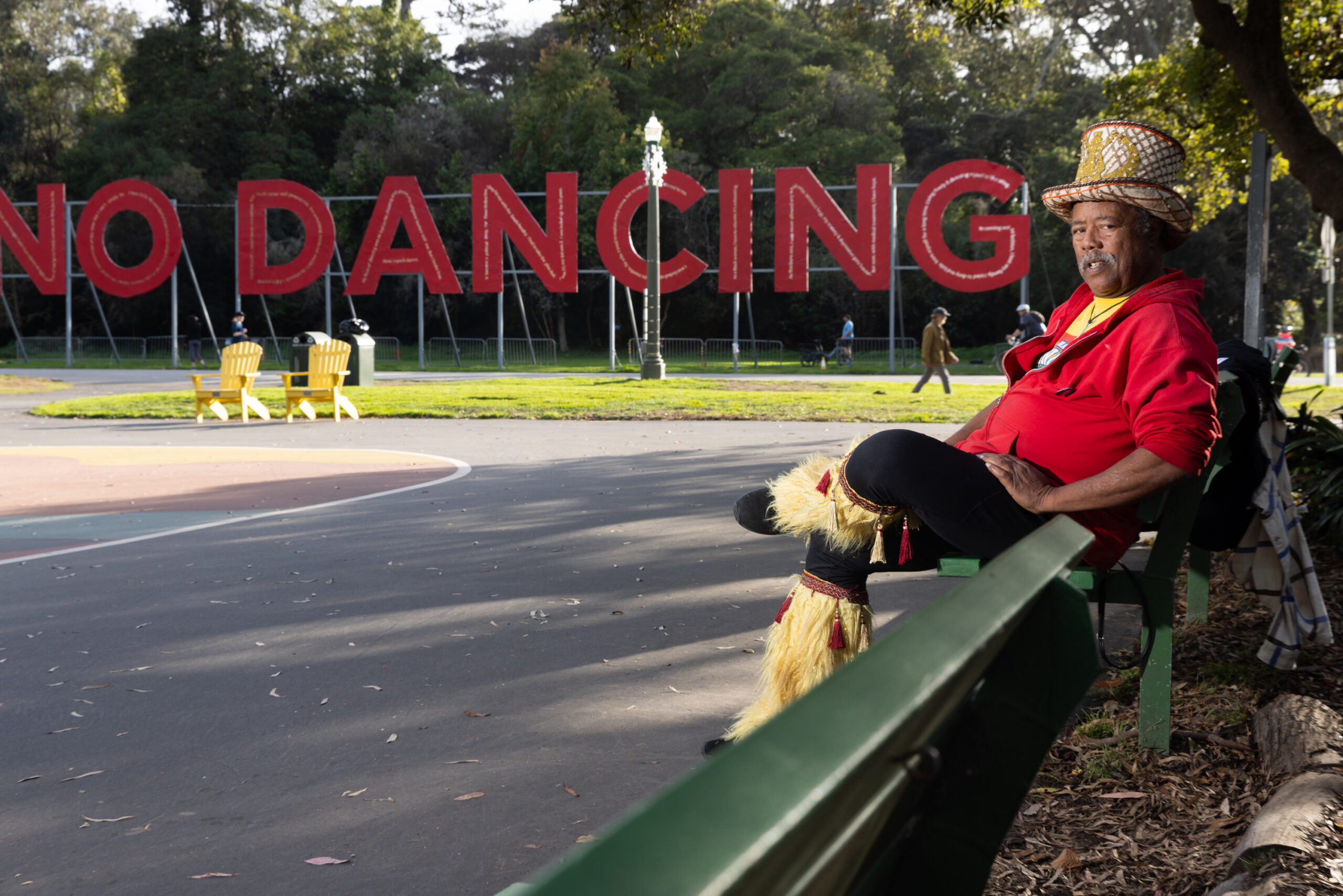 A person with a top hat and red sweater sits on a green bench with a "No Dancing" sign in the back.