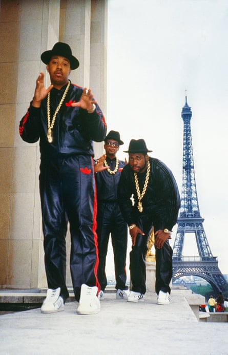 Run DMC in Paris during the Together Forever Tour in 1987. The three are posed with the Eiffel Tower in the background. Run, at left, stands in front, wearing a black and red Adidas track suit, black hat, White and black Adidas shoes, and a large gold chain with medallion. D.M.C., center, is dressed identical to Run, only with a shorter gold chain without a medallion. Both Run and D.M.C. hold their hands up, gesturing at the camera. Jay, at right, wears all black, with a black hat, white and black Adidas shoes, and a large gold chain with medallion. He is bent over with his hands on his knees.