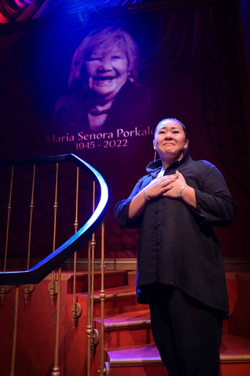 a Filipina woman in black stands on a stage with her hands over her heart in front of a large projected image of her grandmother