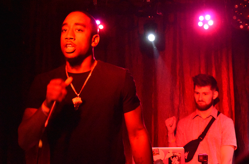 Two individuals are on a stage illuminated by red, blue, and white stage lights. The individual on the left is dressed in a black t-shirt and is holding a microphone. The individual on the right is attired in a white t-shirt and is holding a laptop. The setting suggests a performance scenario.