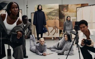 7 people in fancy outfits lounge in front of Italian Renaisance paintings. We see a camera being handed from outside the frame to a Black man kneeling behind a tripod in the bottom right corner.