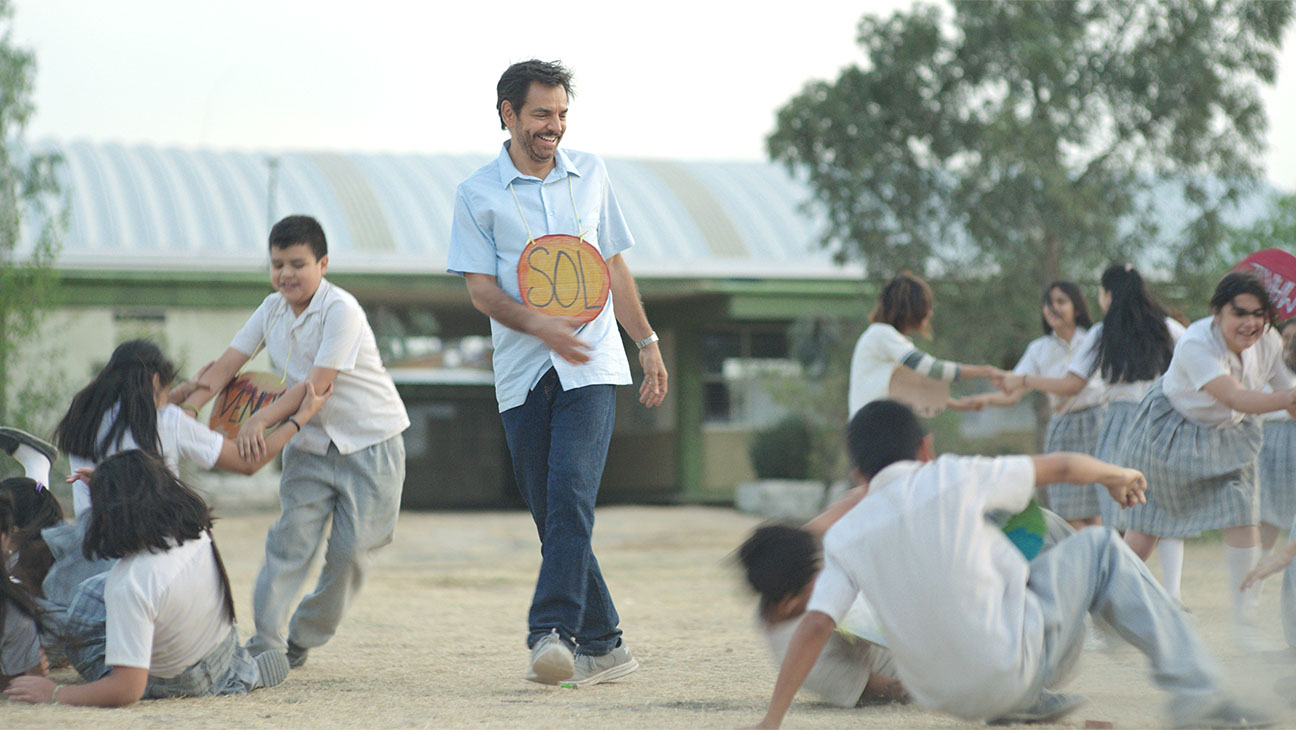 A still from the movie 'Radical' featuring Eugenio Derbez running outdoors with a group of schoolchildren, all of which have a planet in the solar system attached to the front of their shirts